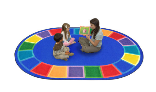 A teacher and two kids sitting on a blue oval rug with seating spaces around the edge of the rug.